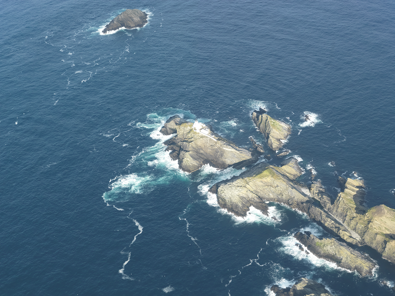 An aerial view of a group of islands with a white lighthouse building on it. A small rocky island lies just above the main group.